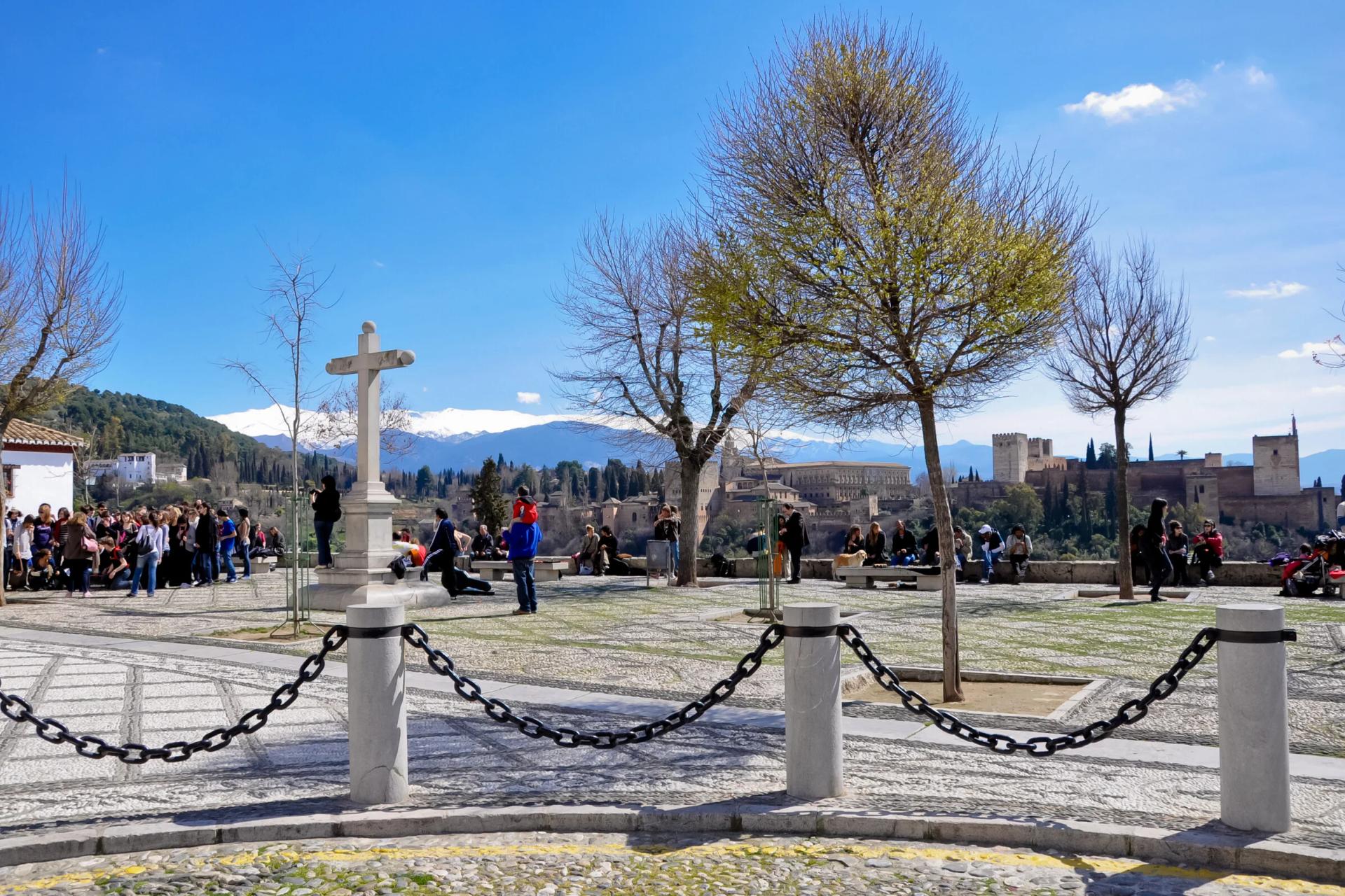 Plaza del mirador de San Nicolás con su cruz de piedra y la gente asomada a la Alhambra y Sierra Nevada