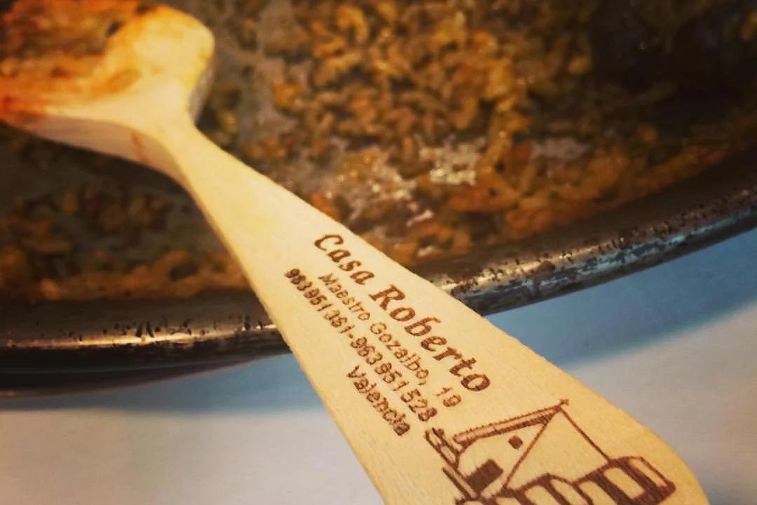 Cuchara de madera con el nombre de Casa Roberto apoyada en una paella